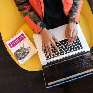 A developer typing code on a laptop with a Python book beside in an office.